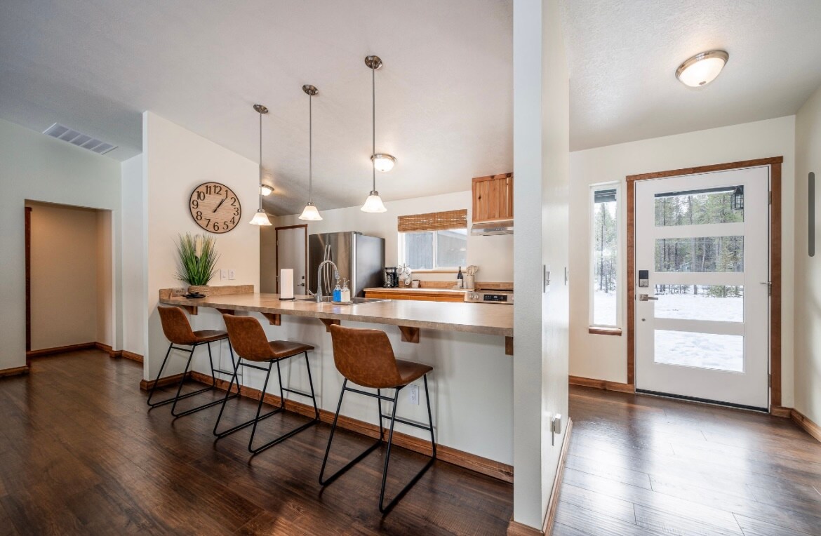 A modern kitchen features a spacious counter with three sleek bar stools. Pendant lights illuminate the area, while a clock decorates the wall. A large window offers outdoor views, and a door leads to the outside, enhancing the open atmosphere.