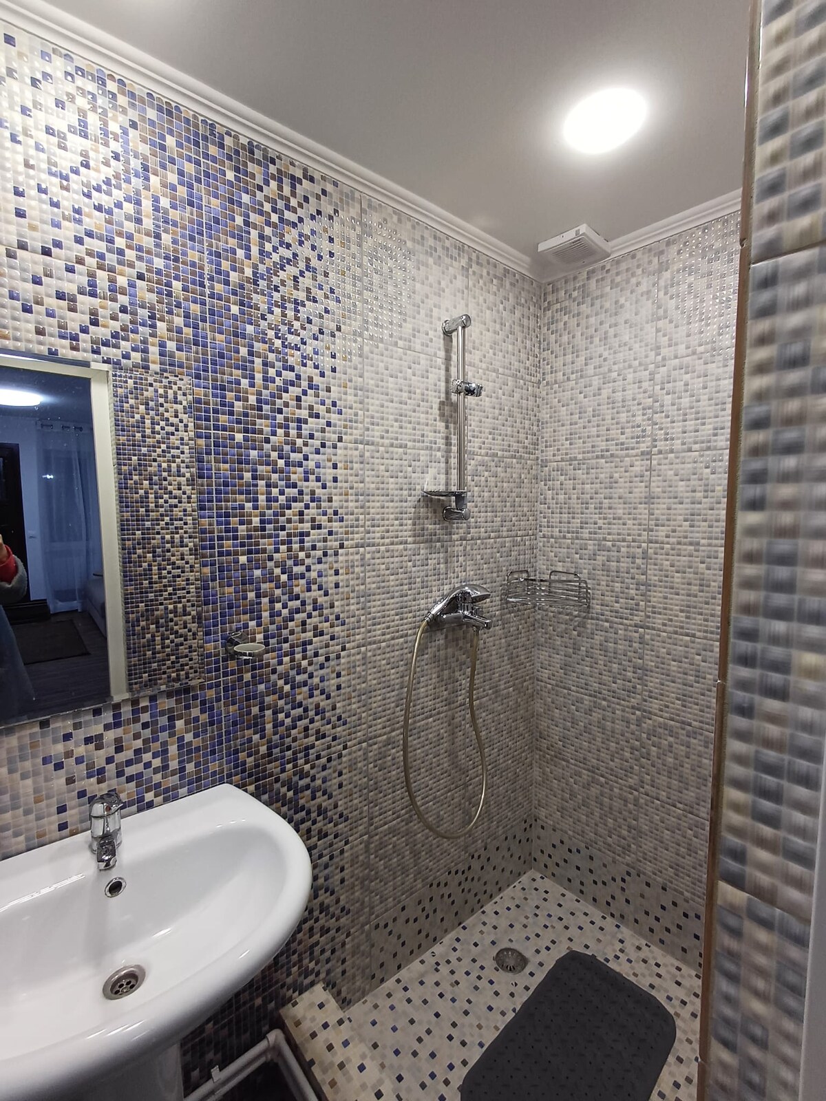A modern bathroom features a walk-in shower with a sleek showerhead and a glass partition. The walls are adorned with small, mosaic tiles in various shades of blue. A compact sink is situated next to a large, framed mirror, enhancing the room's functionality.