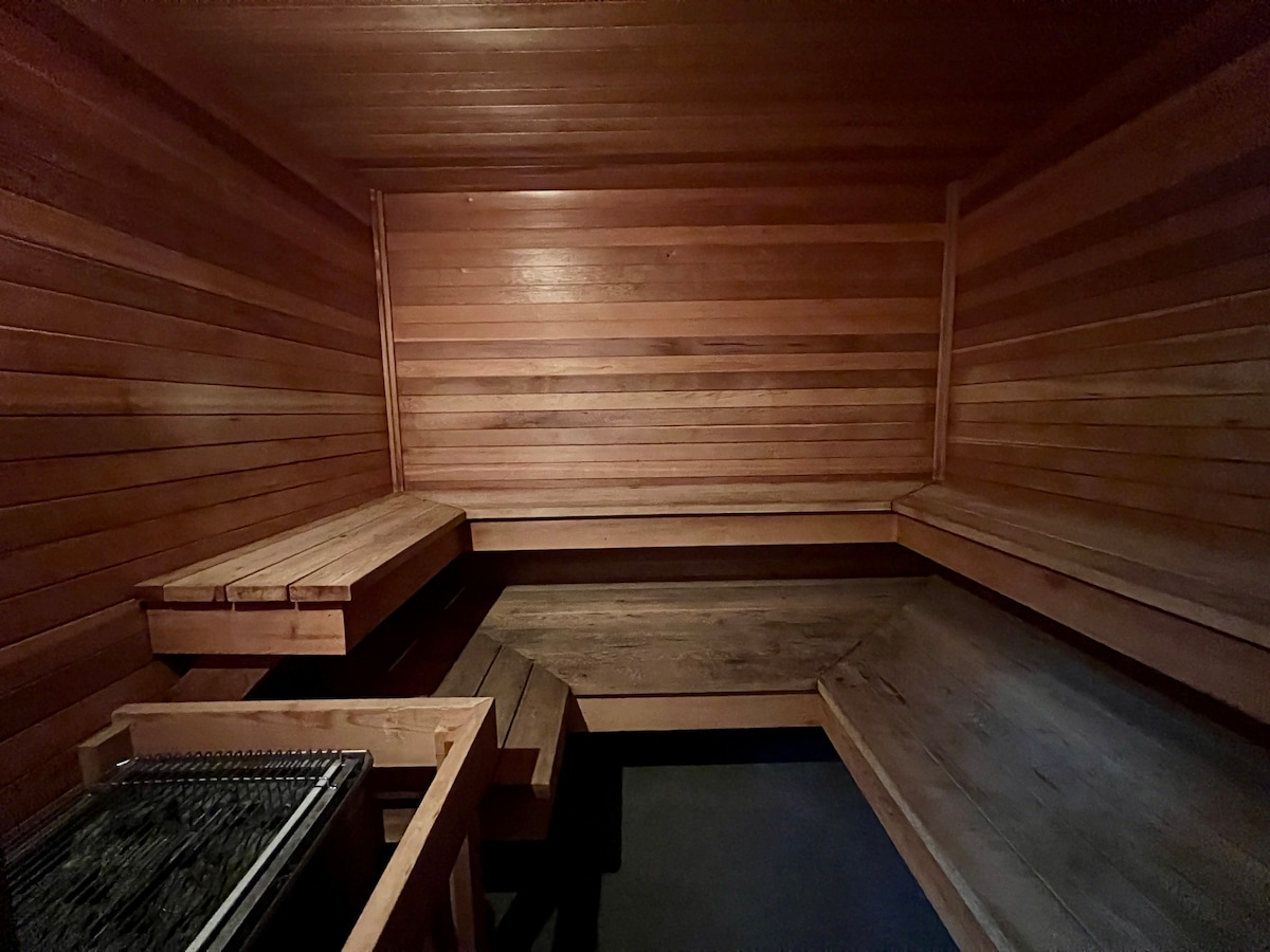 The interior of a wooden sauna is displayed, featuring smooth, warm wooden benches lining the walls. A small wood-burning stove is visible in the corner, contributing to the relaxing atmosphere. The walls showcase a rich, natural wood grain, enhancing the overall serenity.