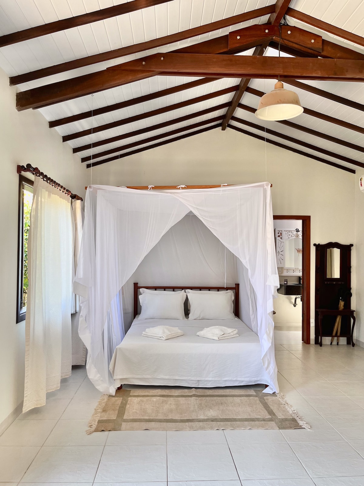 A spacious sleeping area is presented with a wooden bed draped in white fabric. Two neatly arranged white towels are placed on the bed. Natural light enters through large windows, highlighting the high-beamed ceiling and neutral-colored walls.