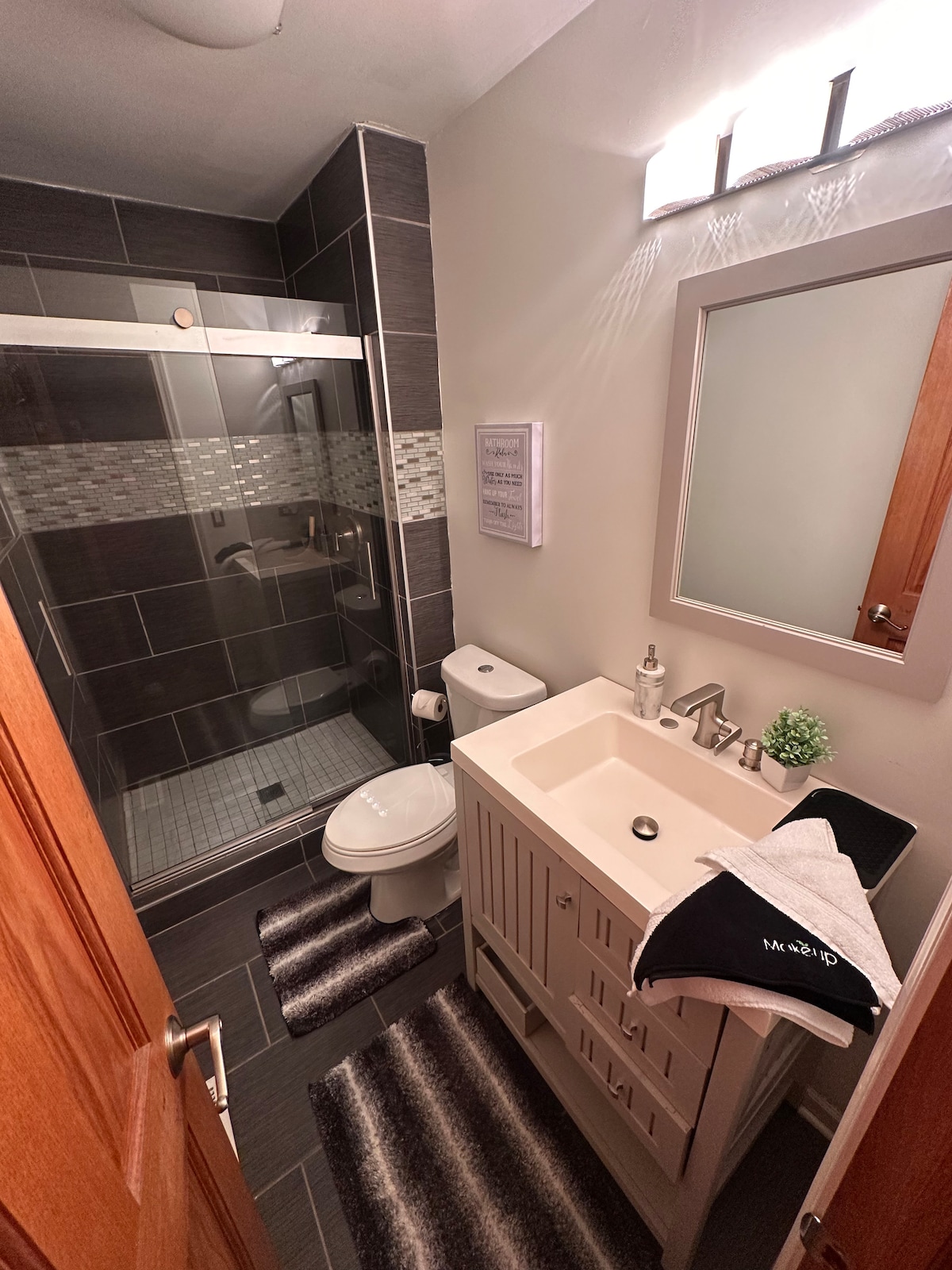 A modern bathroom is presented with dark tiled walls and a glass shower enclosure. A white vanity with a single sink is positioned beside a toilet. Two plush bath mats with a striped pattern are placed on the floor, while a framed mirror reflects the soft lighting above.
