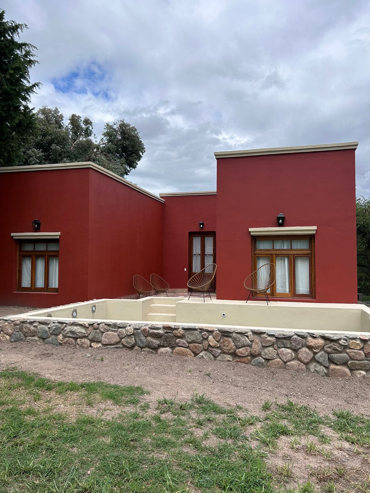 A modern building with a striking red exterior features two spacious windows flanked by light brown wooden accents. The entrance is highlighted by steps leading to a small outdoor area, accompanied by two stylish chairs positioned nearby. Green grass surrounds the property.