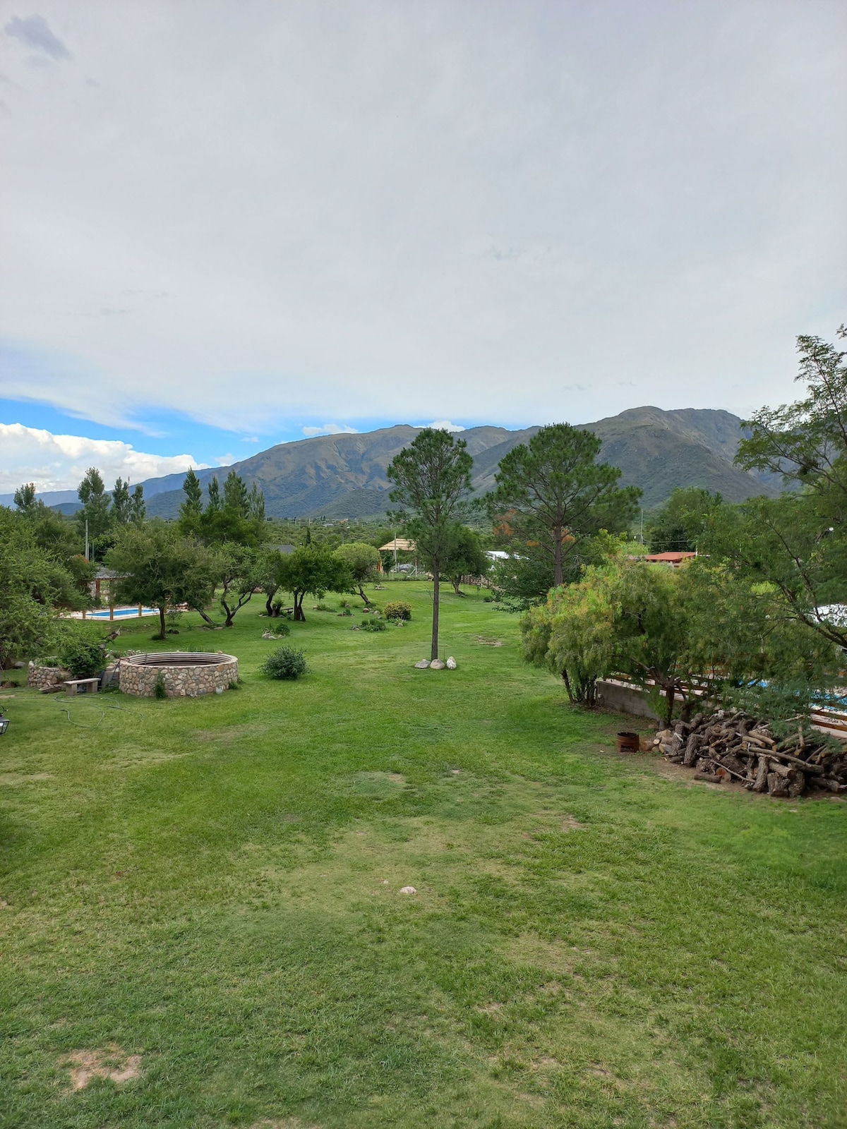 A vast green landscape is framed by distant mountains, featuring a variety of trees and plants. A clear expanse of lawn is visible, alongside a stone structure and firewood stacked neatly. The serenity of the setting is highlighted by the open sky.