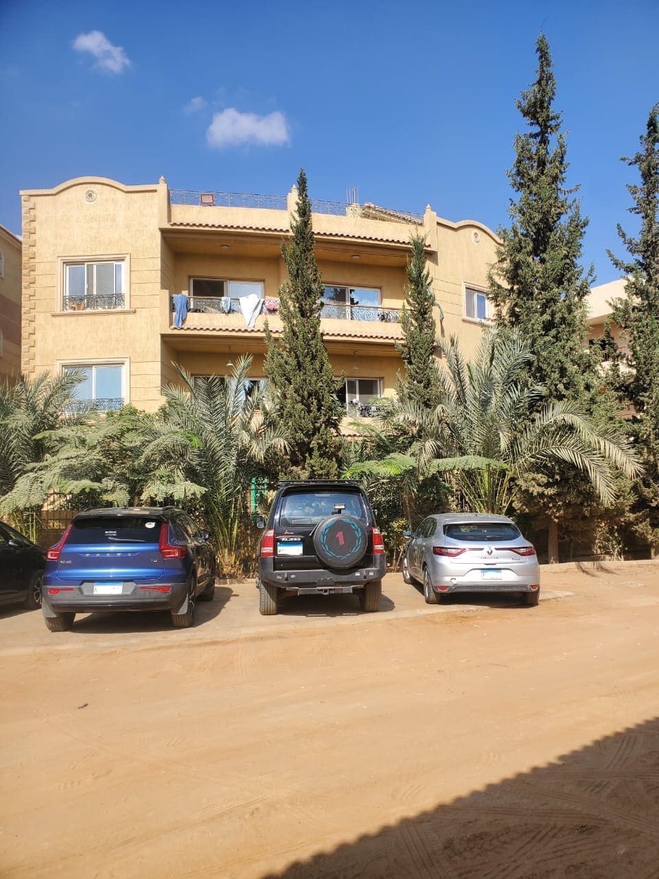 A three-story villa apartment is surrounded by lush greenery, featuring tall conifer trees and ample parking space in front. Vehicles of various sizes are parked on a sandy surface, and open balconies can be seen on the upper floors.