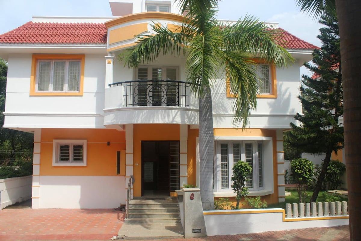 A two-story home features a welcoming entrance with a staircase leading to the door. The exterior showcases a mix of white and orange walls, complemented by a red tiled roof. Lush palms and neatly trimmed shrubbery are visible, enhancing the serene environment.