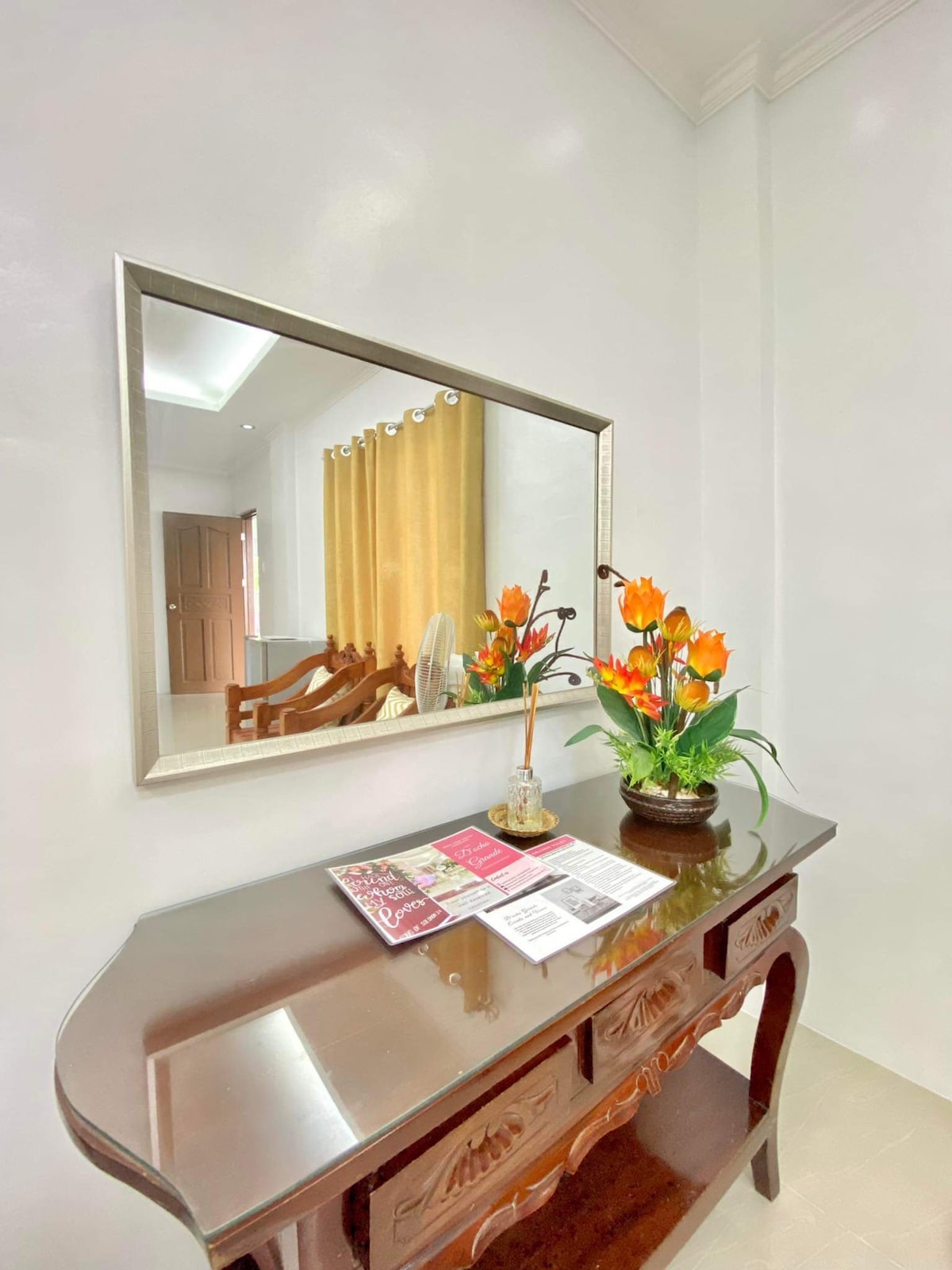 A wooden console table with intricate carvings is positioned against a light-colored wall. A large mirror reflects the room's natural light. A vase of vibrant flowers and a stack of brochures rest on the table, enhancing the welcoming ambiance.