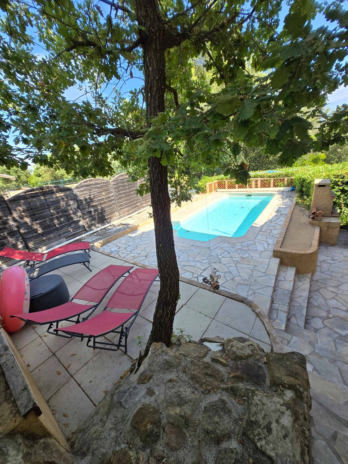 A tranquil outdoor area features a swimming pool surrounded by stone paving. Several lounge chairs are positioned beside the pool, and a tree provides shade. Lush greenery encloses the space, creating a private and peaceful atmosphere.