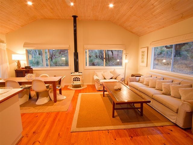 A cozy living area is highlighted by a plush sofa, a wooden coffee table, and a dining space with modern chairs. Large windows provide ample light and views of the surrounding nature. A wood stove adds warmth to the inviting atmosphere.