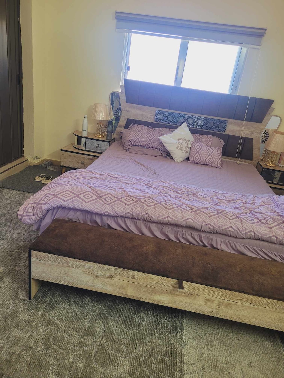 A spacious bedroom features a wooden bed frame with a padded headboard and a light purple bedding set. A decorative pillow is placed on the bed. Light enters through a window, complemented by a small bedside table with lamps.