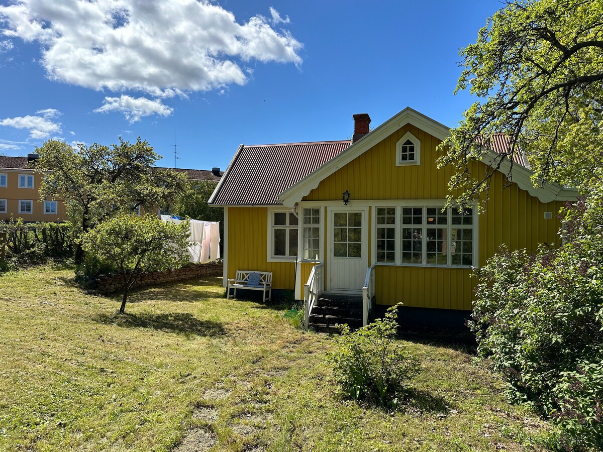 A yellow main building is visible, set in a spacious garden surrounded by trees and bushes. The structure features a welcoming front porch and multiple windows, allowing natural light to brighten the interior. The scene is complemented by a clear blue sky and scattered clouds.