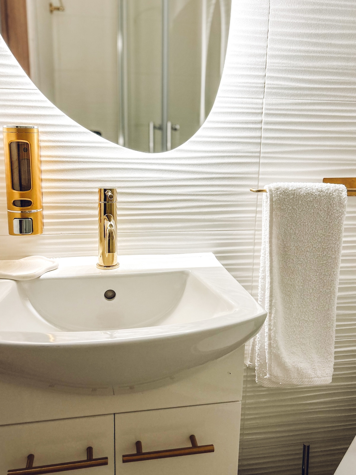 The bathroom features a modern sink with a stylish faucet and a soap dispenser mounted on the wall. A neatly hung white towel is displayed beside the sink. The area is illuminated by natural light reflecting off a round mirror, enhancing the overall brightness.