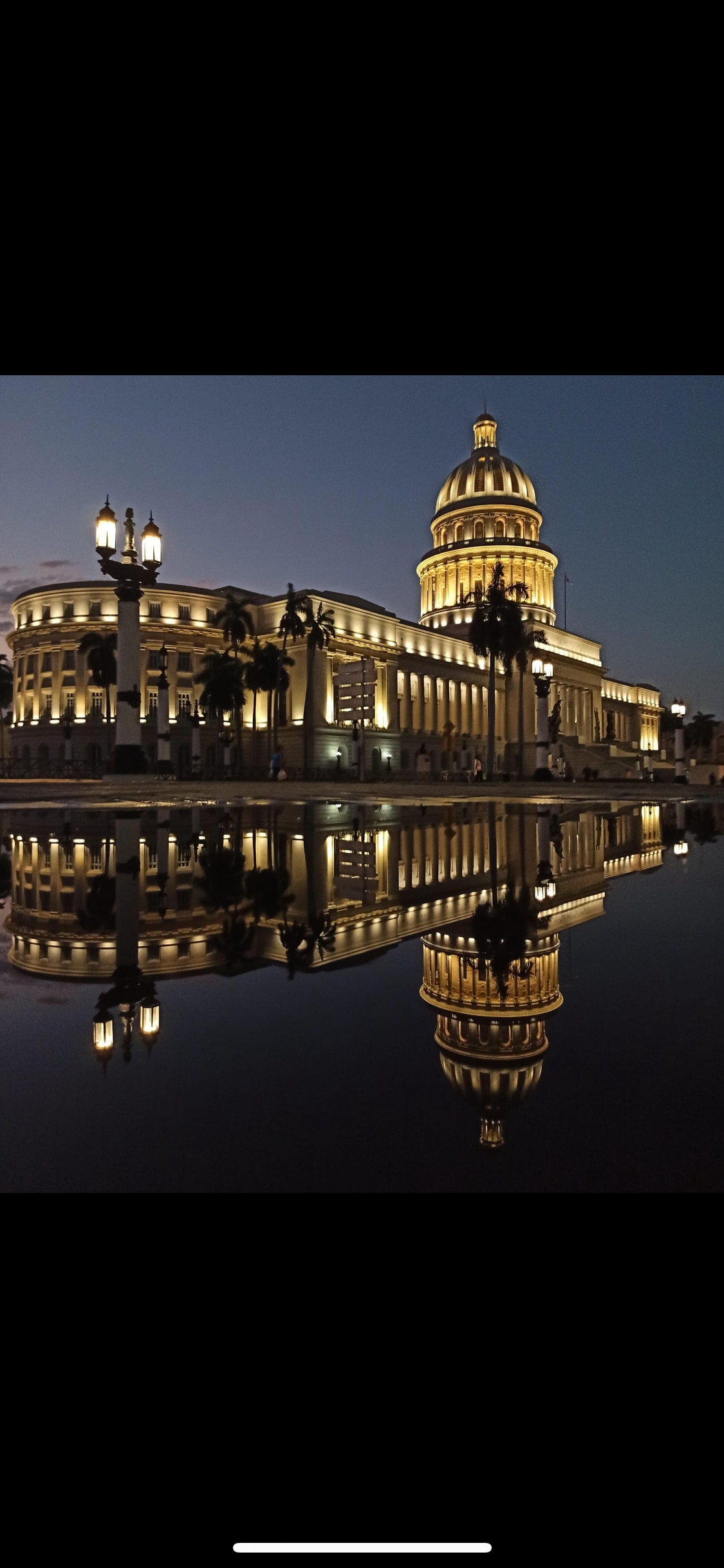 The Capitolio building is illuminated against a twilight sky, showcasing its grand architectural details. The structure is reflected in a still body of water in the foreground, enhancing its impressive stature. Soft lights highlight the building's dome and ornate facade.