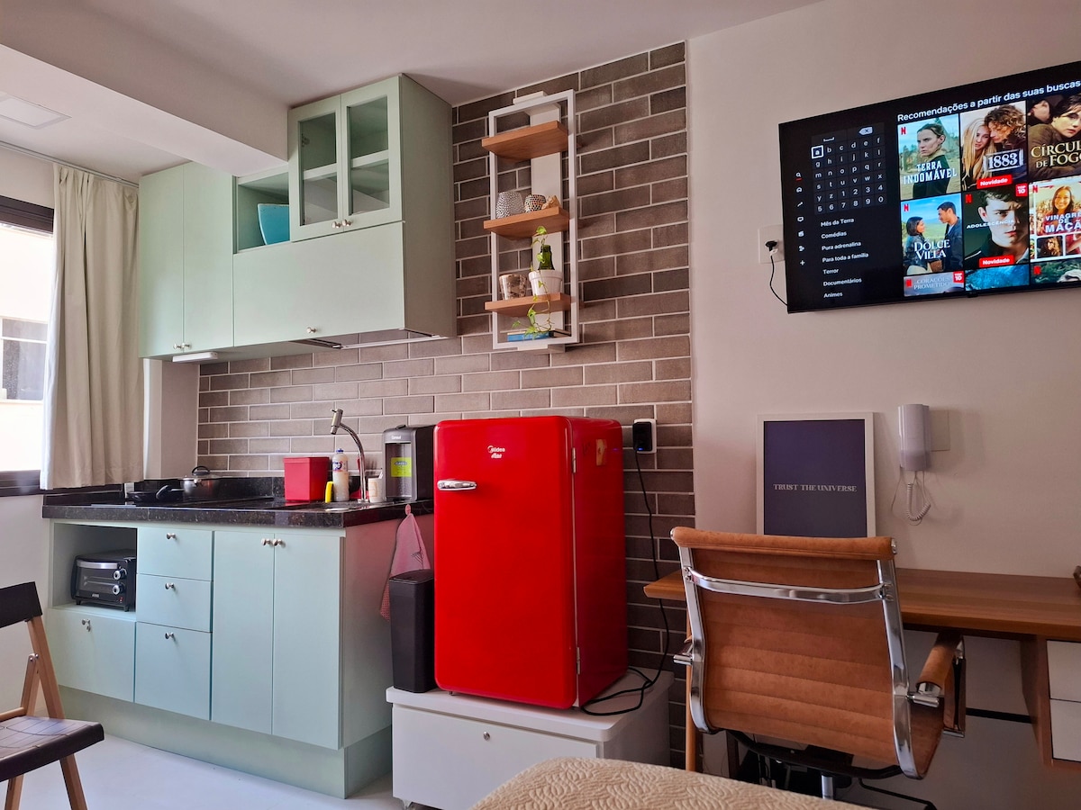 A functional kitchenette features sleek cabinetry in soft green hues, complemented by a dark countertop. A vibrant red refrigerator stands out against the neutral tile backdrop. A mounted TV displays various streaming options, while a wooden desk with a chair provides a workspace. Natural light enters through a window.