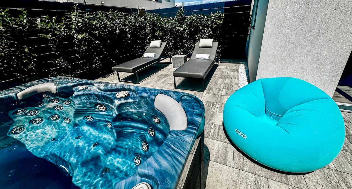 An outdoor area features a hydromassage hot tub with clear blue water, surrounded by stone tiles. Two lounge chairs are placed nearby, alongside a large, bright blue bean bag chair. Lush greenery provides privacy on the perimeter.