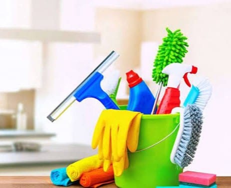 A green bucket filled with various cleaning supplies is displayed on a countertop. Items include an orange scrub brush, bright yellow gloves, a blue squeegee, spray bottles in red and blue, and multi-colored cleaning cloths, providing a sense of cleanliness and preparation.