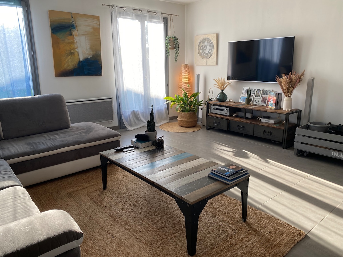 A spacious living area features a large sectional sofa and a rustic coffee table, adding character to the space. Natural light filters through two large windows, illuminating decor elements like a potted plant and wall art. A TV is mounted on the wall, enhancing the cozy atmosphere.