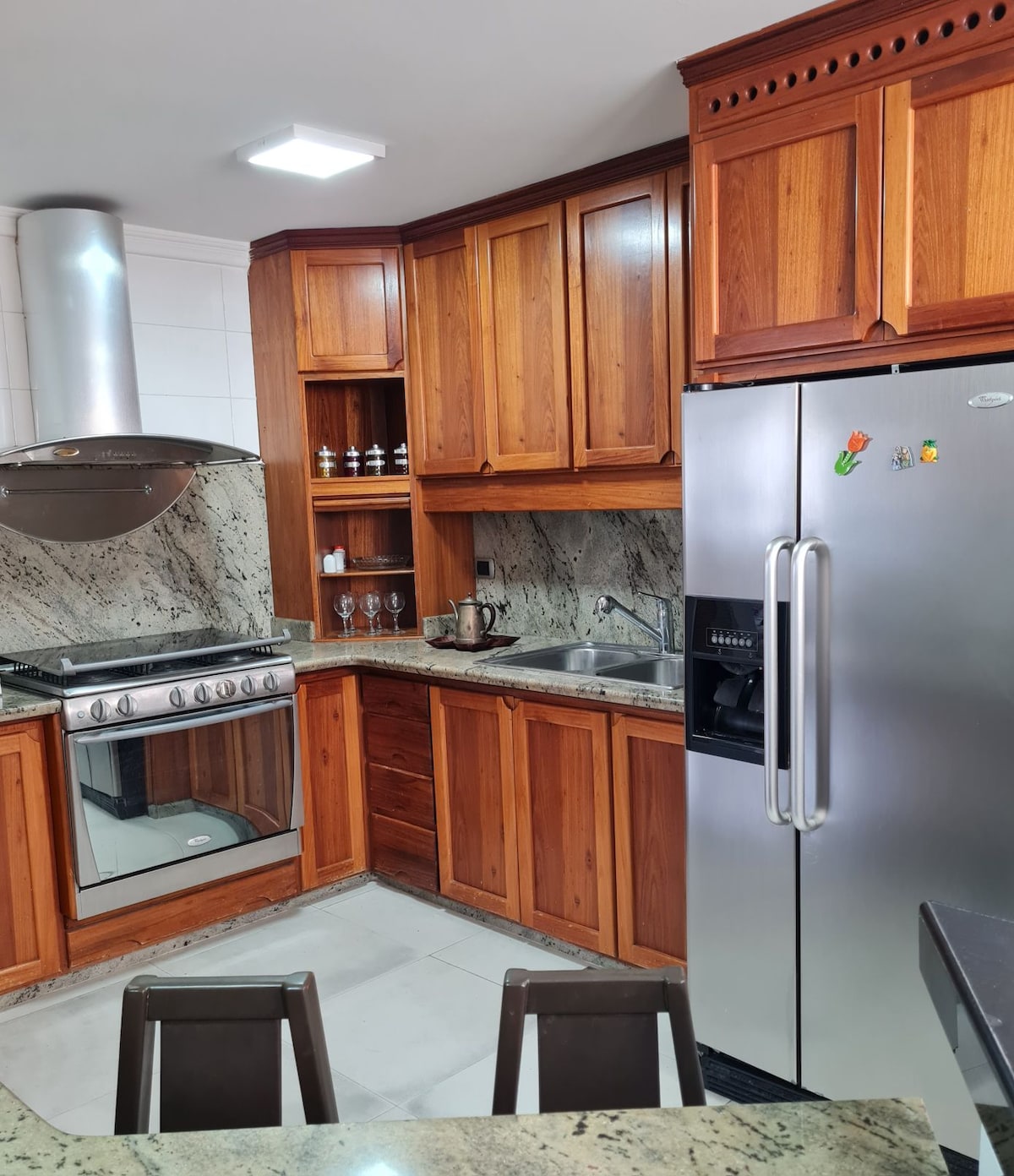 A welcoming kitchen features warm wooden cabinetry complemented by a granite countertop. Stainless steel appliances, including a refrigerator and a gas stove, are present. Glassware is displayed on a nearby shelf, and ample counter space is provided for meal preparation.