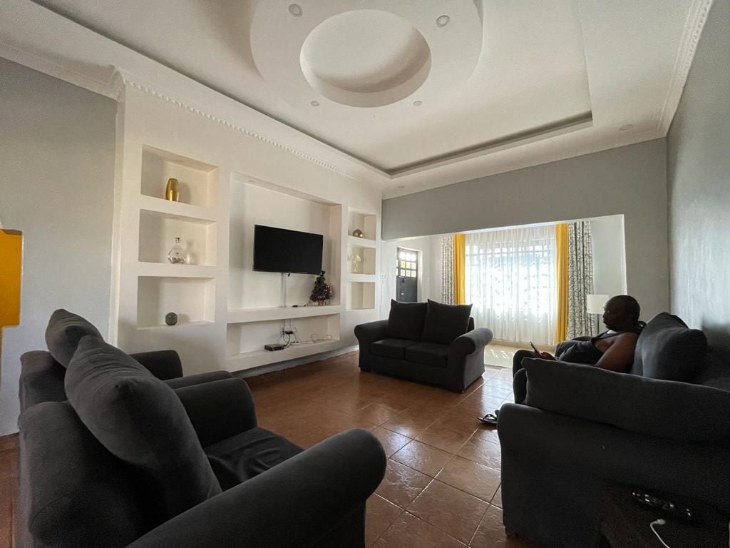A spacious living area is furnished with two comfortable gray sofas positioned for relaxation. A large flat-screen television is mounted on a white wall unit, complemented by decorative shelves. Natural light filters through sheer curtains, creating a bright and inviting atmosphere.