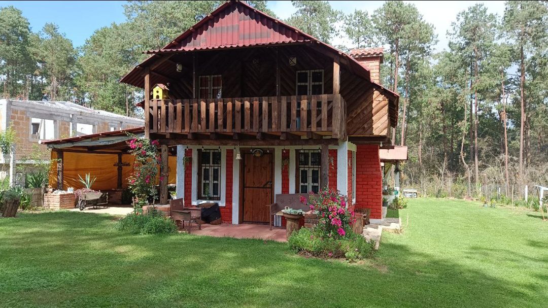 A charming cabin built with natural materials is nestled in a lush green setting. The two-story structure features a wooden balcony, vibrant flowers, and comfortable seating on a stone patio. A well-maintained lawn surrounds the cabin, offering a serene outdoor space.