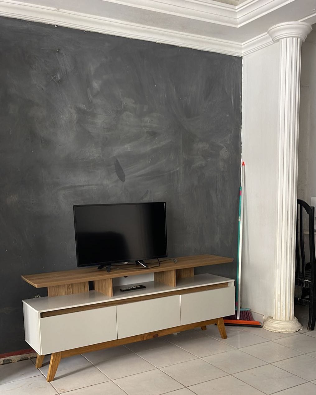 A sleek television is mounted on a wooden stand, positioned against a dark feature wall. Below the TV, a storage unit with white drawers provides space for media items. A broom is seen leaning against the wall to the right, and a column adds architectural character to the room.