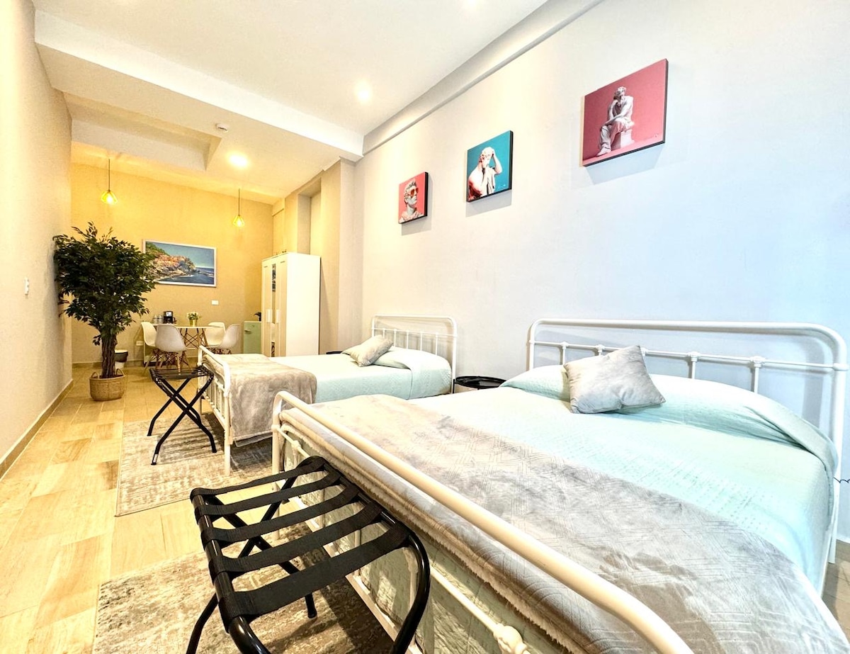 A bright, airy space features two spacious beds covered with pastel bedding. A decorative wall showcases three framed artworks. A small dining table with chairs is positioned in the corner, and a potted plant adds a touch of greenery to the room.