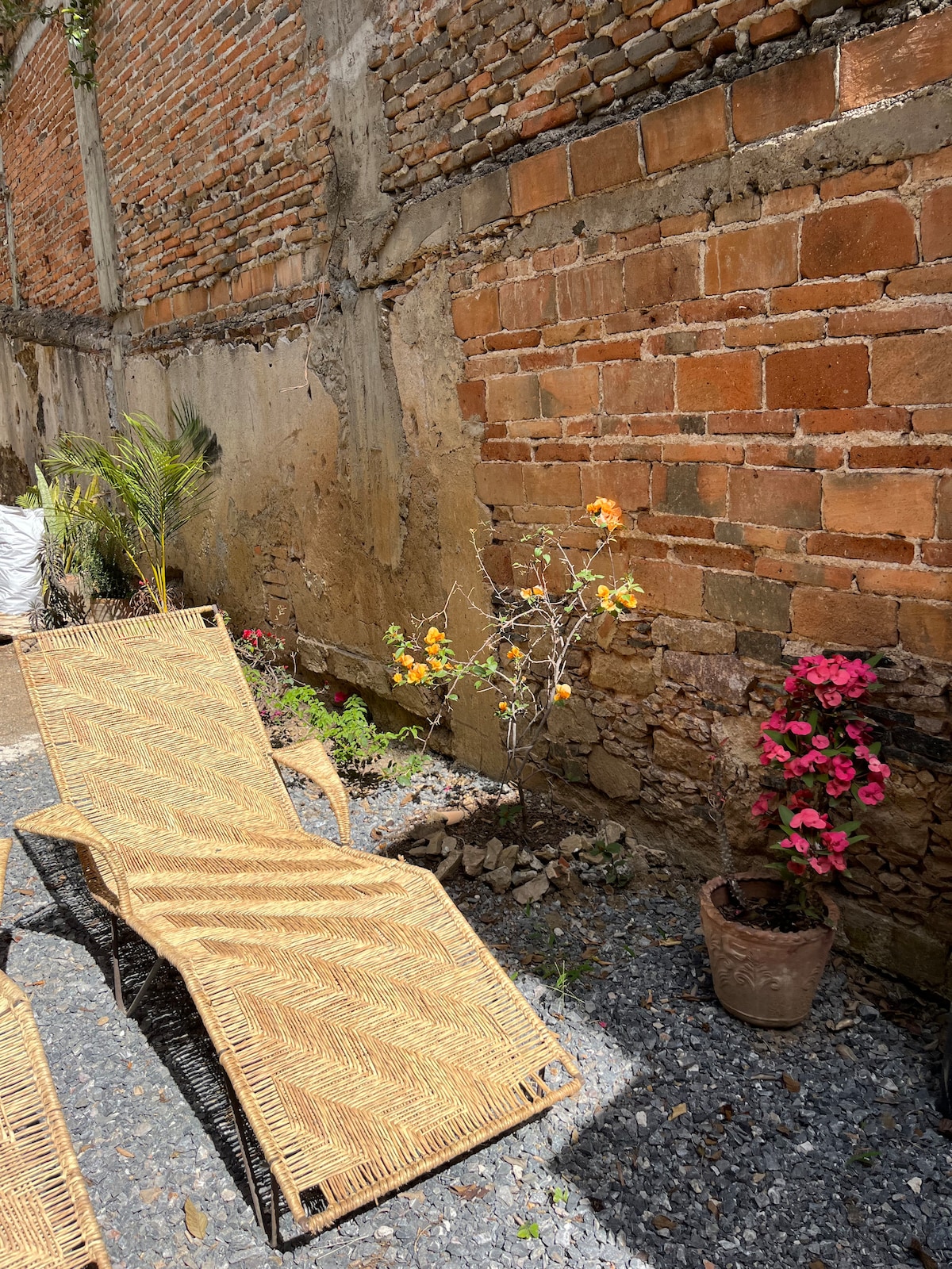 Two sun loungers are positioned on a gravel surface, surrounded by a rustic wall made of exposed brick. Potted plants with blooming flowers add a touch of color to the space, creating a serene outdoor area for relaxation.