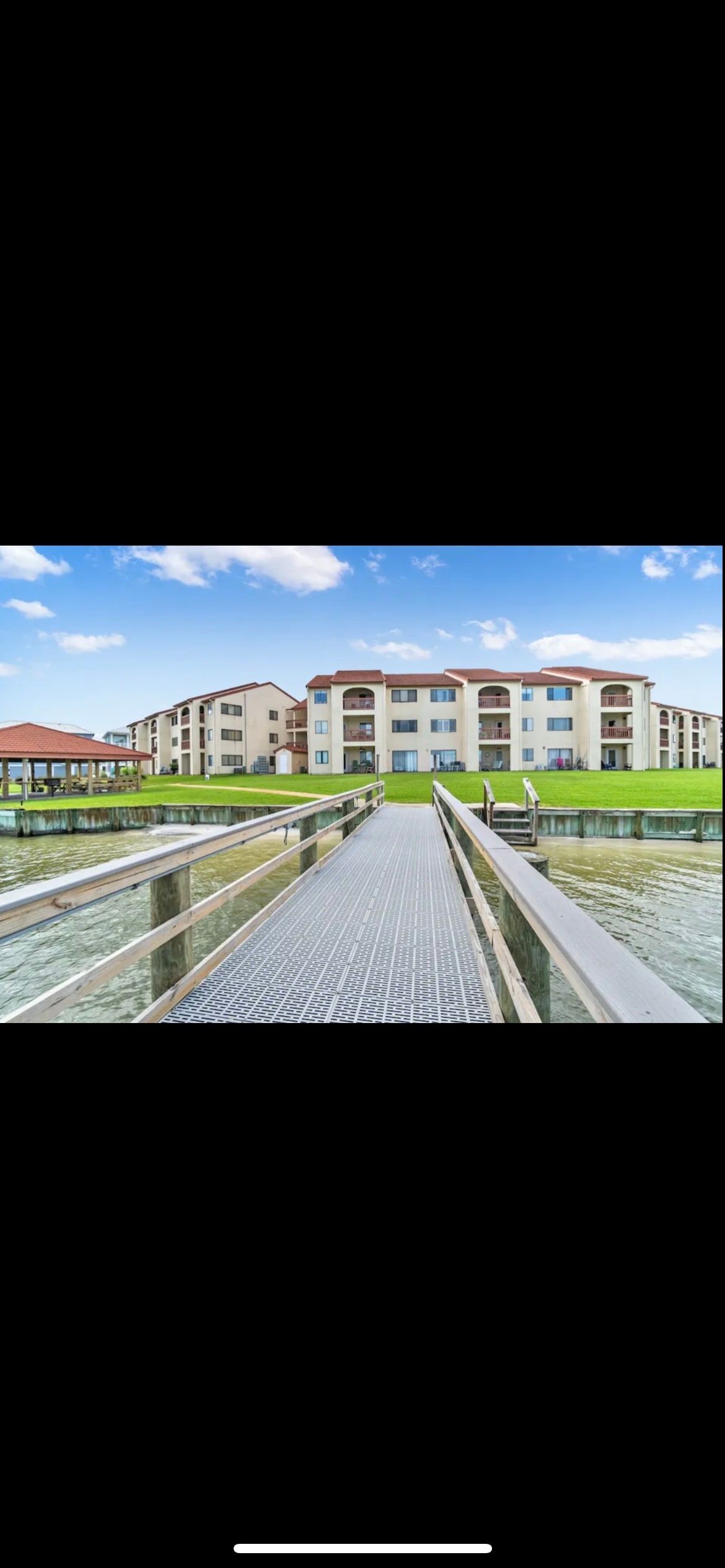 A view from the pier shows a walkway leading towards a modern condo building. The structure features multiple balconies and large windows, reflecting a serene waterfront environment with lush green grass lining the edges.