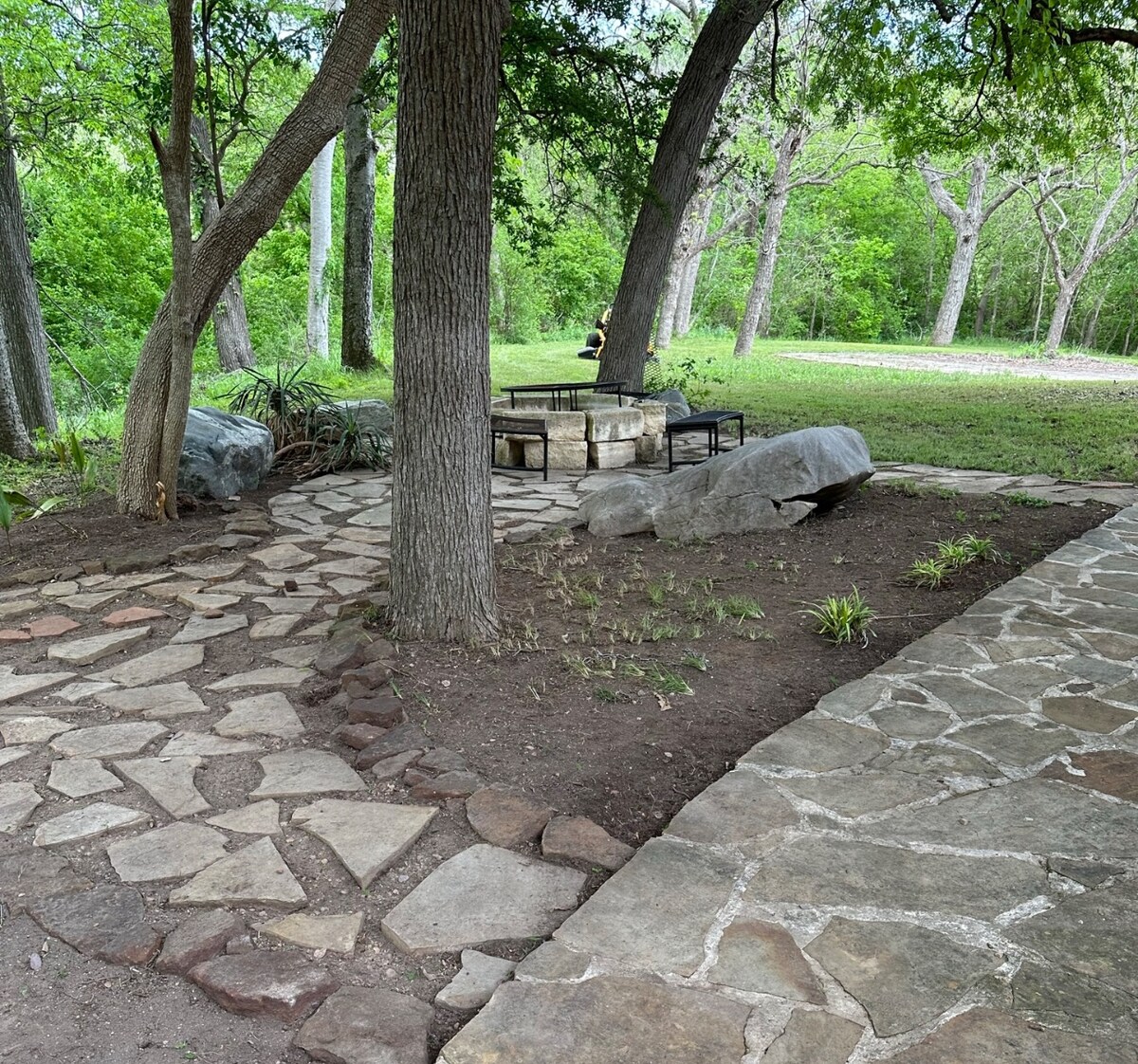 A stone pathway meanders through a shaded area featuring large trees and natural landscaping. A rustic fire pit surrounded by seating stones offers an inviting space for gatherings, while lush greenery provides a serene backdrop.