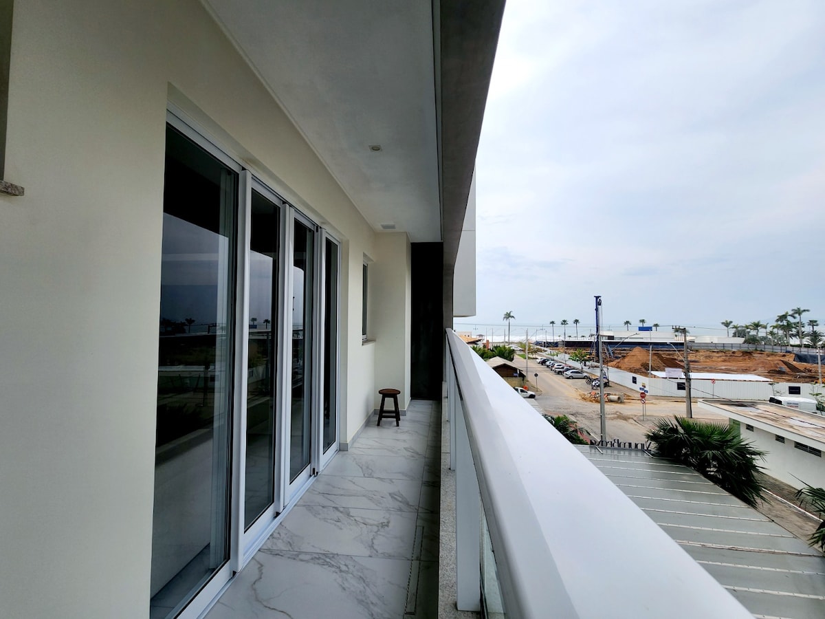 A spacious balcony features smooth marble flooring and large glass sliding doors, offering views of the surrounding area. A single wooden stool is positioned near the entrance, and the horizon is visible in the distance, showcasing the proximity to the beach.
