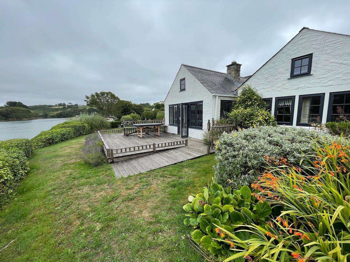 A waterside cottage is presented, showcasing an outdoor deck area with seating. Lush greenery and blooming flowers frame the space, while the peaceful creek and distant landscape provide a serene backdrop under an overcast sky.