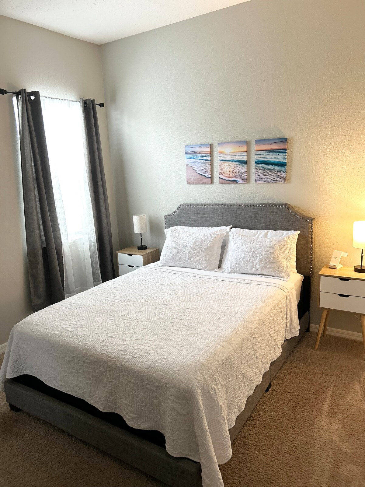 A well-appointed sleeping space features a queen bed dressed in white linens. Artwork depicting serene beach scenes is displayed above the bed. Two bedside tables with lamps flank the bed, and natural light filters through the nearby window adorned with gray curtains.