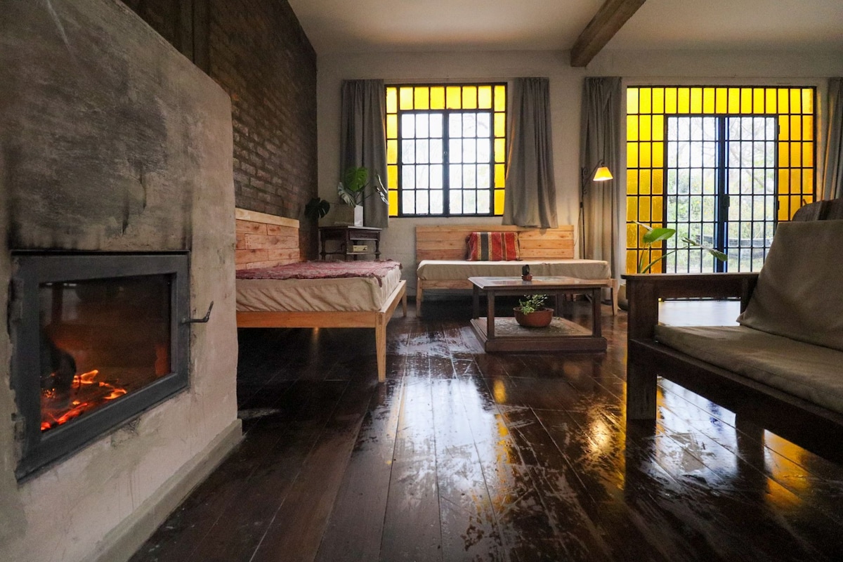 An open space living area features a cozy fireplace and seating arrangements with natural light streaming through large, yellow-tinted windows. Historic wooden flooring complements the modern industrial design, while a small table with a potted plant adds a touch of greenery to the environment.