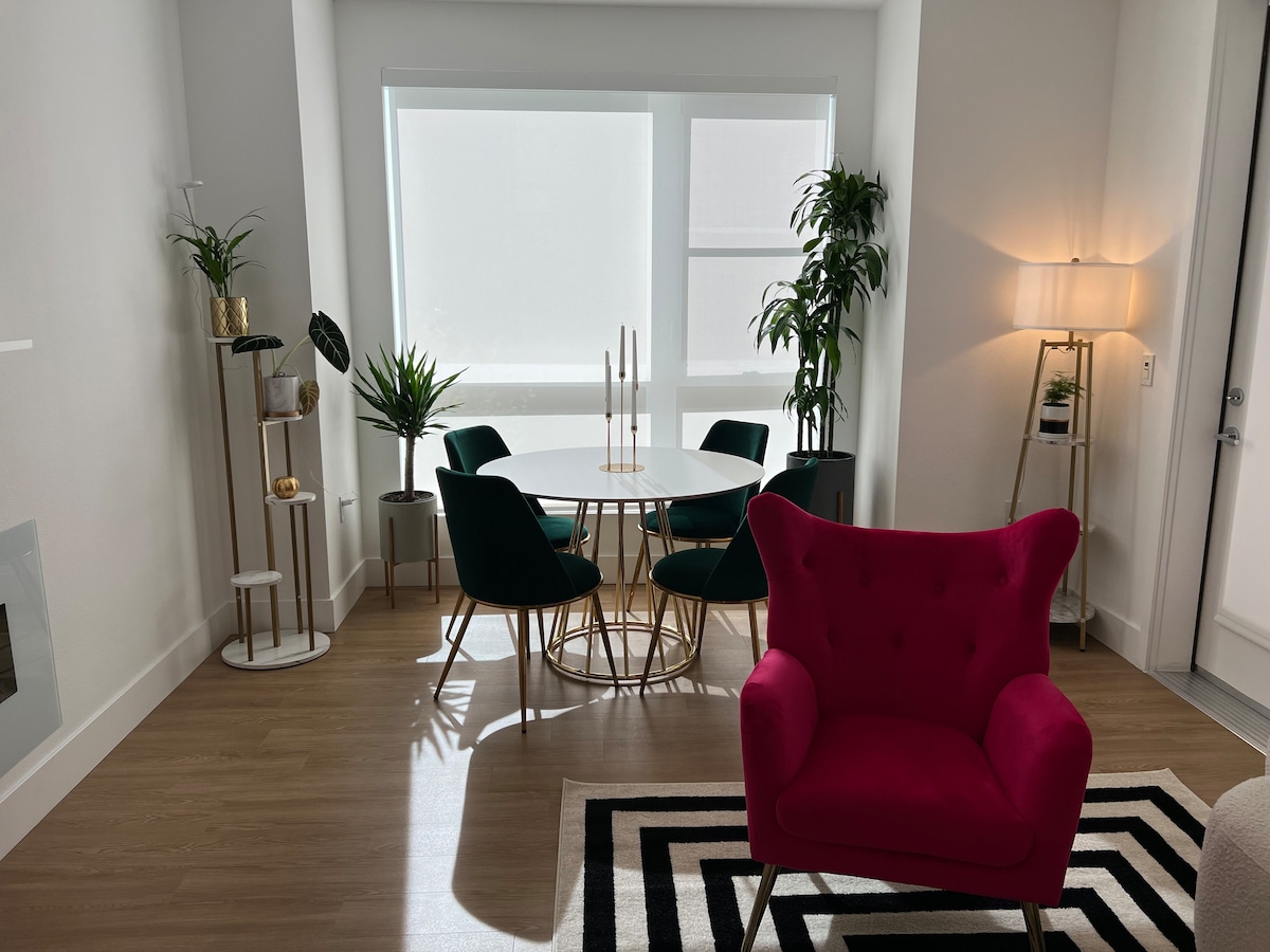 A stylish dining area is visible, featuring a round table surrounded by dark green upholstered chairs. A tall, slim floor lamp stands beside a decorative plant. Light filters through large windows, and a vibrant pink accent chair adds a pop of color to the space.