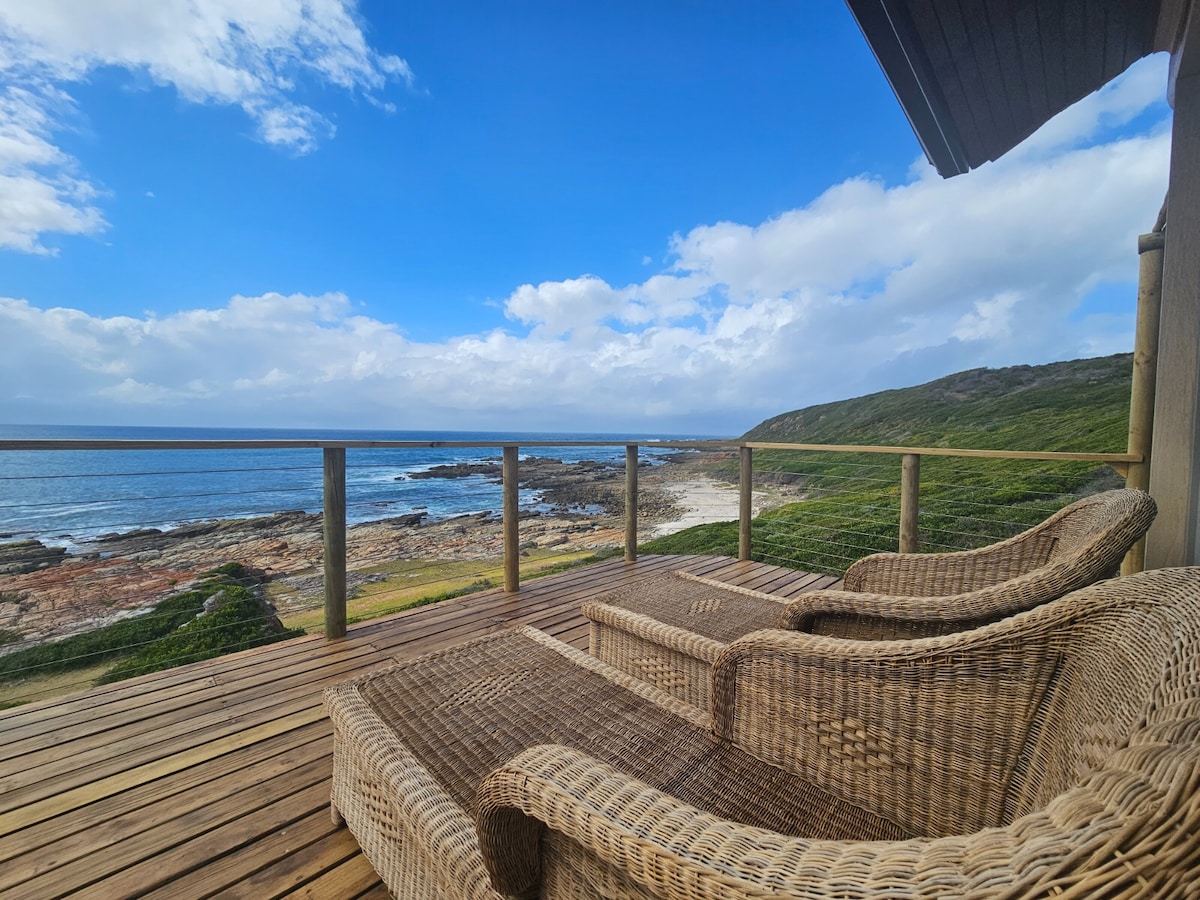 A spacious balcony features two woven loungers overlooking the ocean and rocky coastline. The calm sea reflects the blue sky with scattered clouds, while greenery is visible in the background, creating a serene coastal setting.