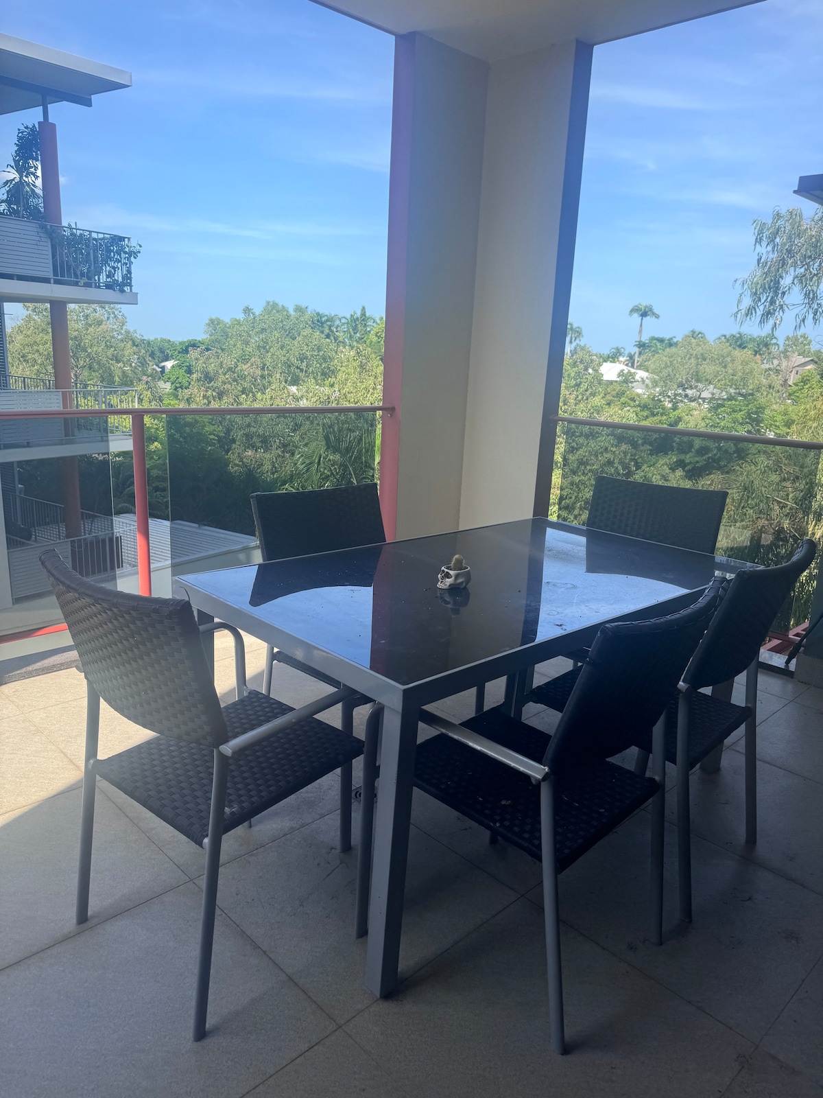A dining area features a glass-top table surrounded by six black wicker-style chairs. The space is shaded by a balcony overhang, with lush green trees visible in the background. Natural light brightens the area, creating a refreshing atmosphere.