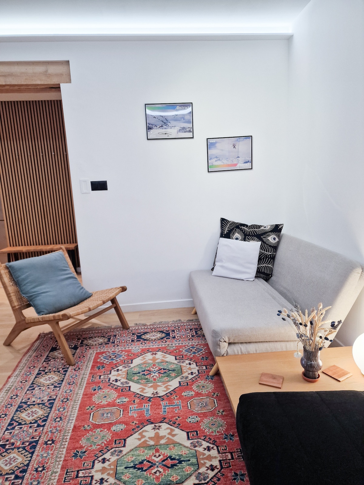 A modern living area is presented with a light-colored sofa bed, accompanied by a woven chair. Decorative wall art adorns the space, while a patterned area rug adds color. A wooden table holds a vase with dried flowers, and soft light is provided by a lamp.