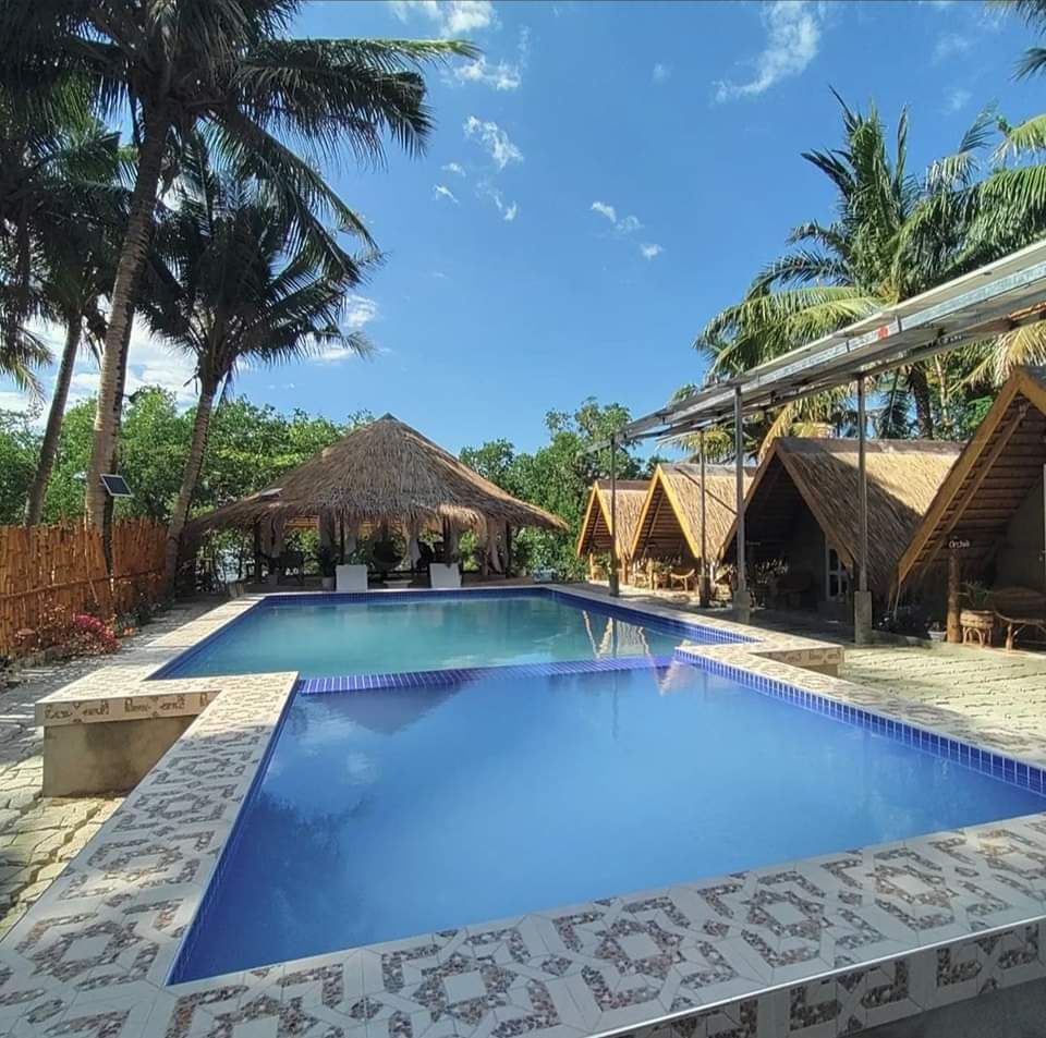 A clear blue swimming pool is framed by palm trees and thatched-roof huts. Lounge chairs are placed around the pool area, providing ample space for relaxation. The bright sky enhances the tranquil ambiance of the outdoor setting.