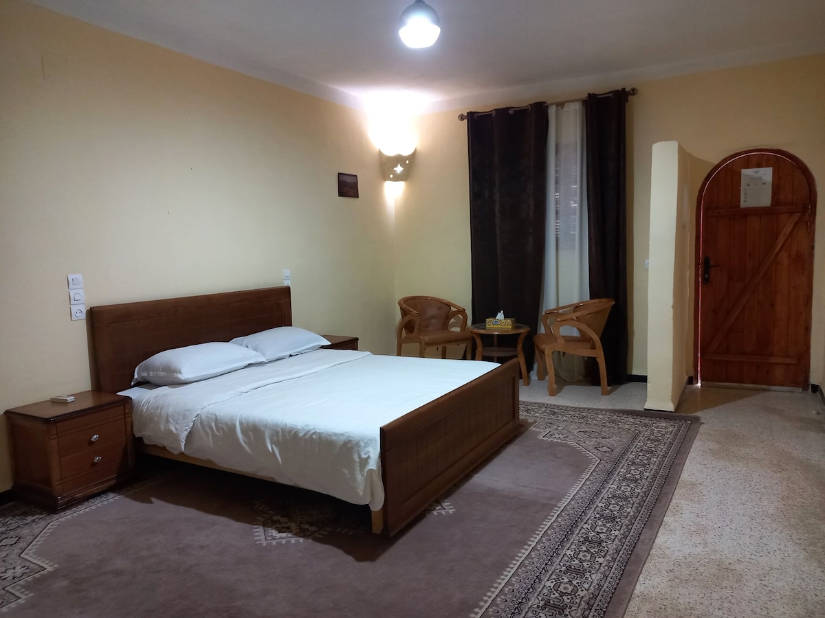 A simple bedroom features a spacious bed with a white blanket, positioned against a light-colored wall. Two wooden chairs are situated beside a decorative rug, and a door with wooden framing leads to another area, while soft lighting enhances the space's warmth.