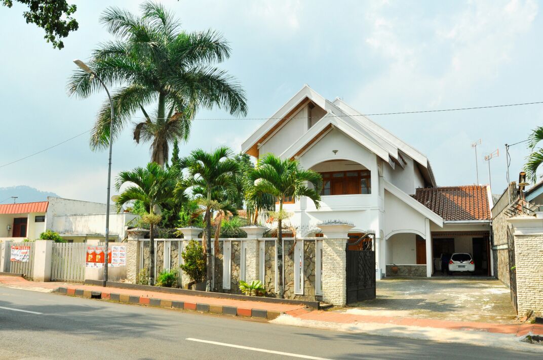 A spacious two-story house is surrounded by greenery, featuring palm trees in the front yard. A stone wall with a gated entrance provides privacy, while a driveway leads to an area for parking. The architecture is modern and bright, enhancing the inviting exterior.