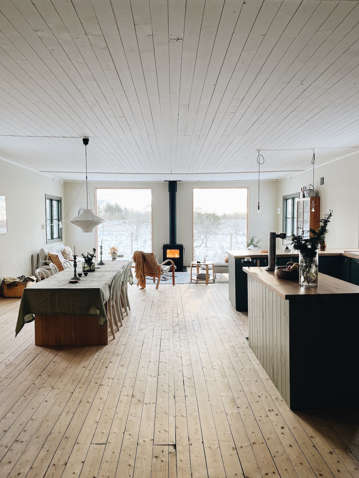 An open living space features a long dining table with seating for several people, complemented by a cozy seating area near a wood-burning stove. Natural light streams in through large windows, illuminating the wooden flooring and neutral-colored walls.