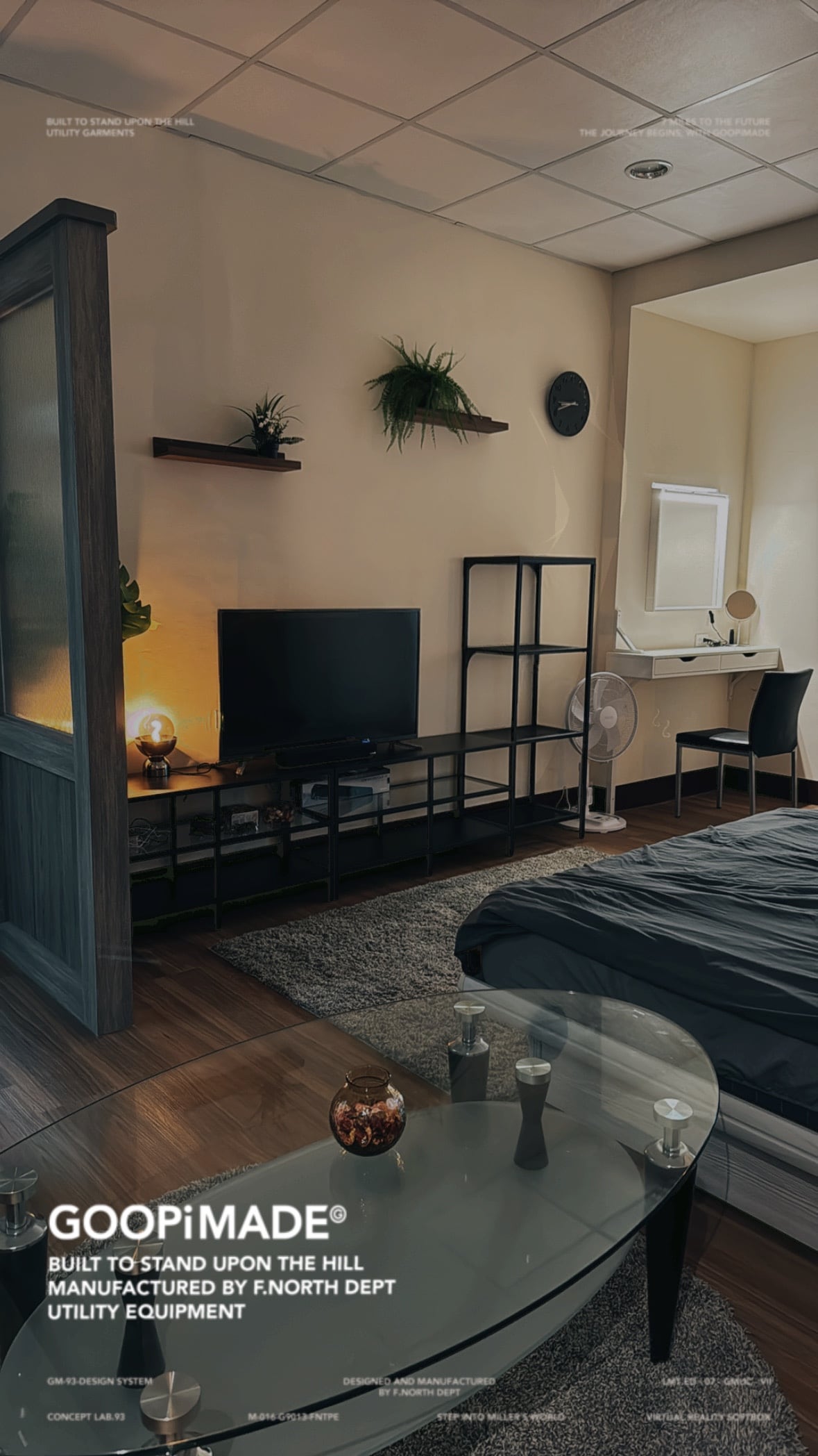 A modern living space is presented with a spacious bed dressed in dark linens. A glass table is positioned in front of a low, sleek TV unit. A desk and chair are visible against the wall, alongside a standing fan and a wall clock, creating a functional environment.