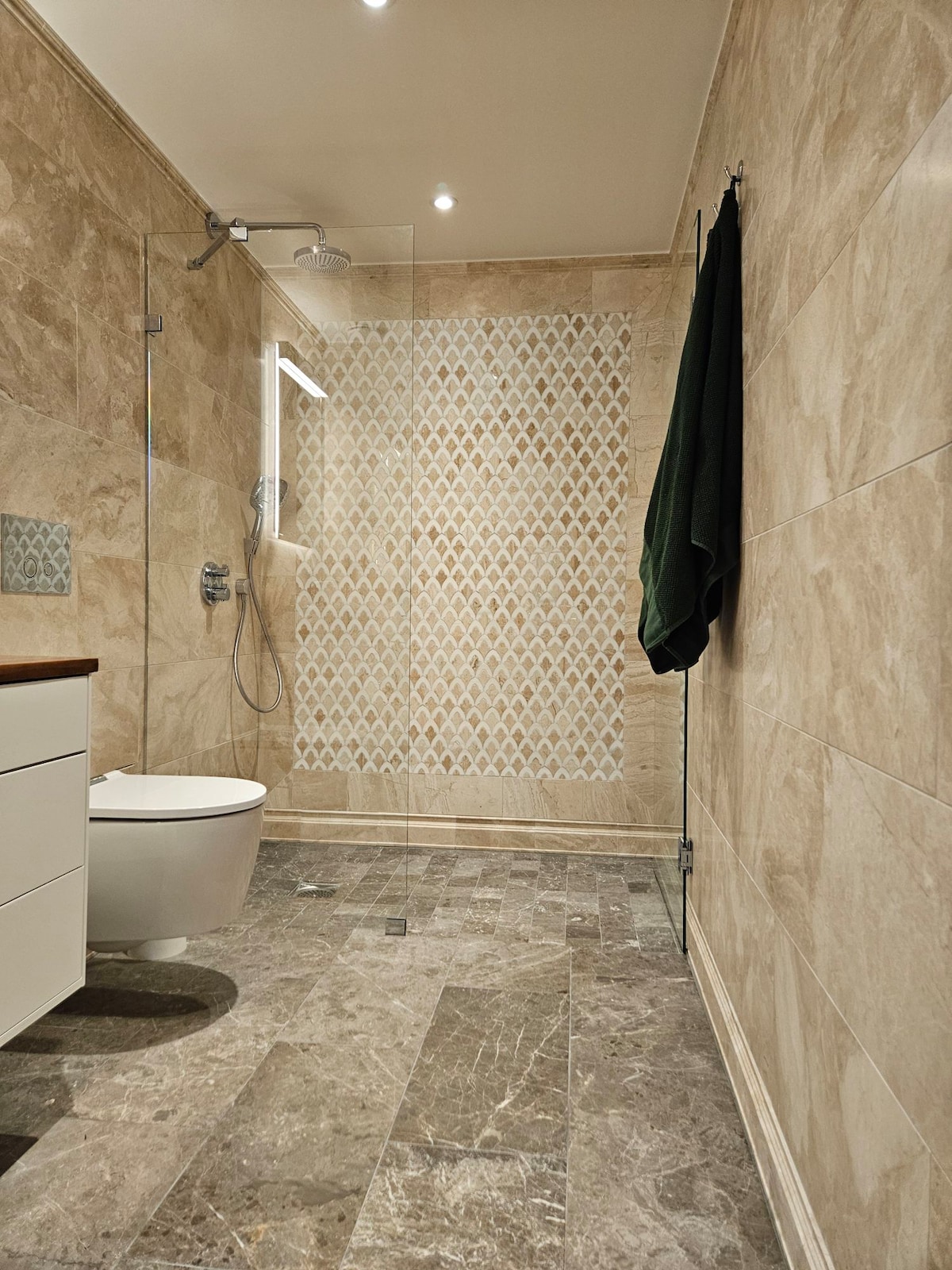 A modern bathroom features marble walls and flooring, creating a sleek appearance. A glass shower enclosure is equipped with an overhead shower, while a stylish light fixture provides illumination. A dark green towel adds a touch of color, and a white toilet is discreetly positioned in the corner.