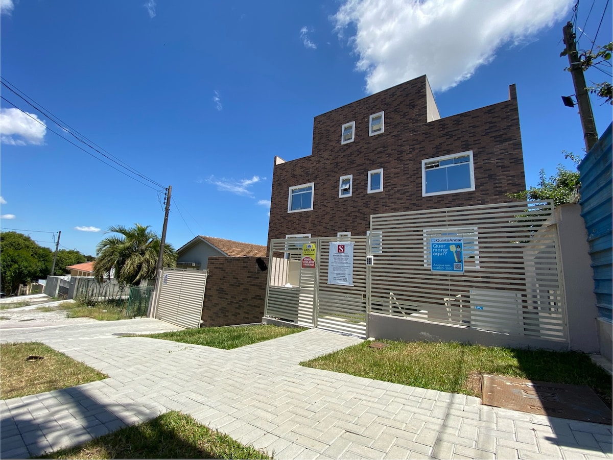 A modern building with a distinctive brick facade is situated amidst green grass under a clear blue sky. A paved pathway leads to the entrance, framed by decorative fencing. Surrounding areas feature well-maintained landscaping and an open space that offers easy access.