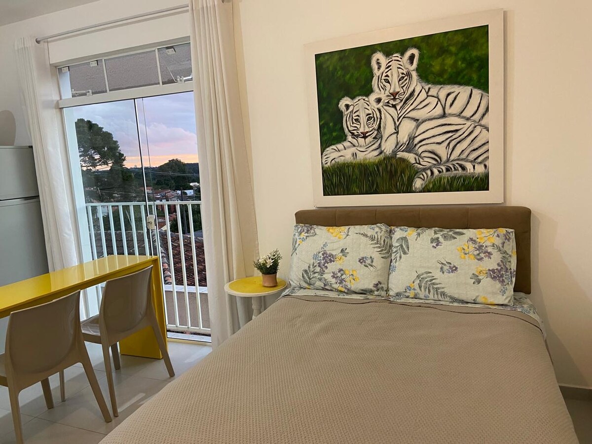 A spacious bed is adorned with a floral-patterned duvet, complemented by a neutral-colored throw. A vibrant painting of two white tigers hangs above the bed. Nearby, a small round table is set against a window with a view of the outdoors, featuring two chairs.
