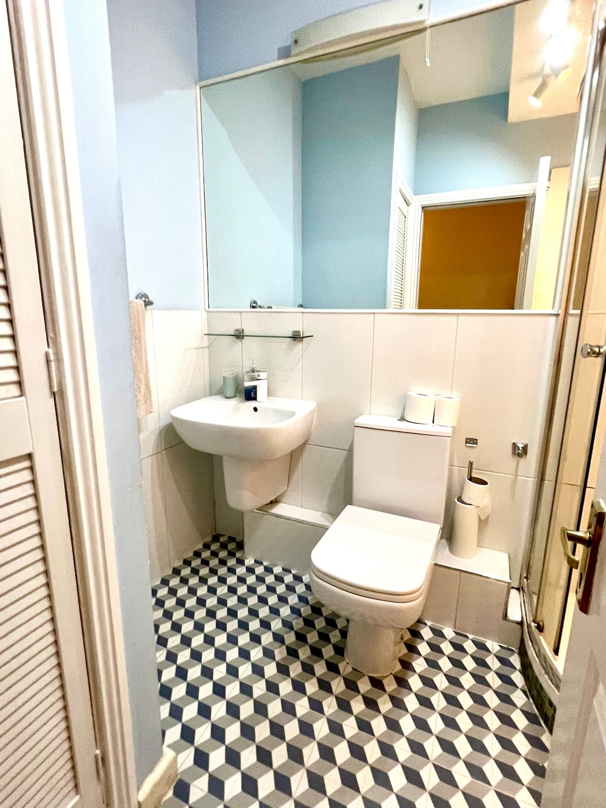 The bathroom features a modern design with white tiled walls and a geometric patterned floor. A glass shower stands alongside a sink and toilet. A large mirror above the sink reflects the space, enhanced by natural light from the doorway.