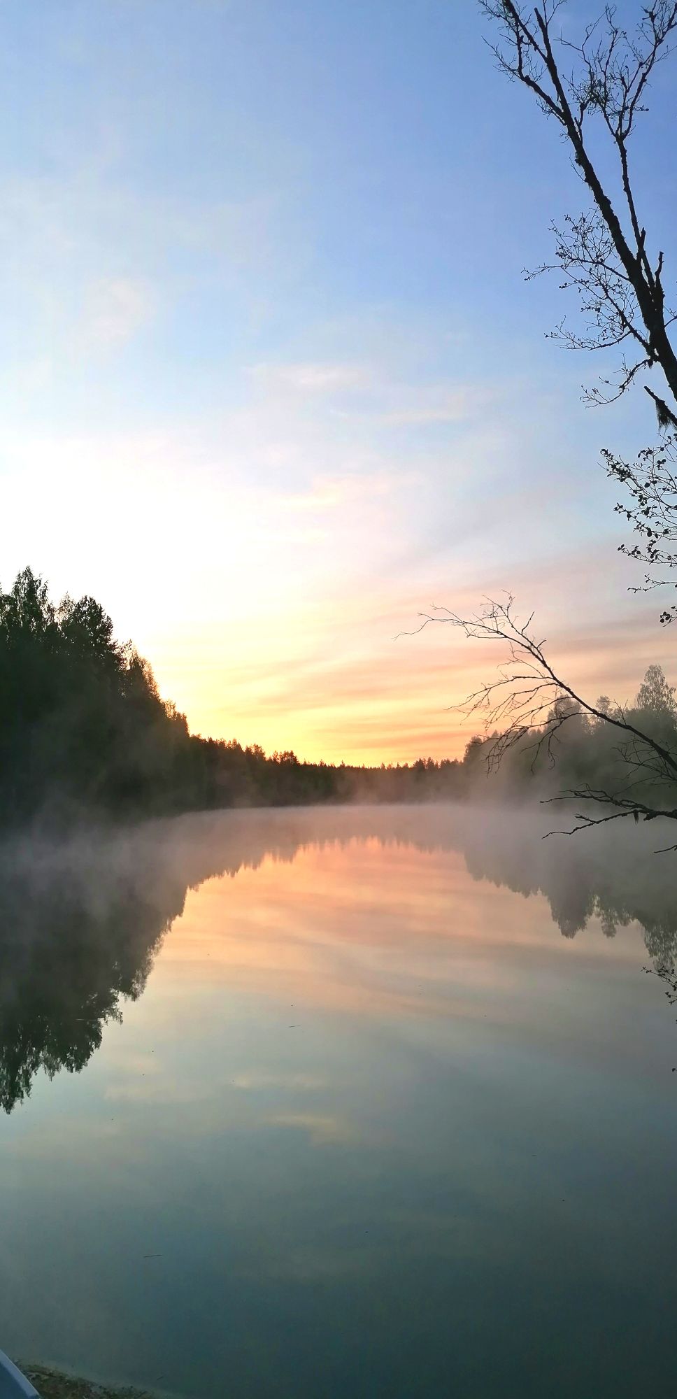 A calm river reflects soft hues of orange and blue during sunrise, surrounded by tall trees. A light mist hovers above the water’s surface, creating a serene atmosphere. The scene conveys a sense of tranquility, inviting relaxation in a natural setting.