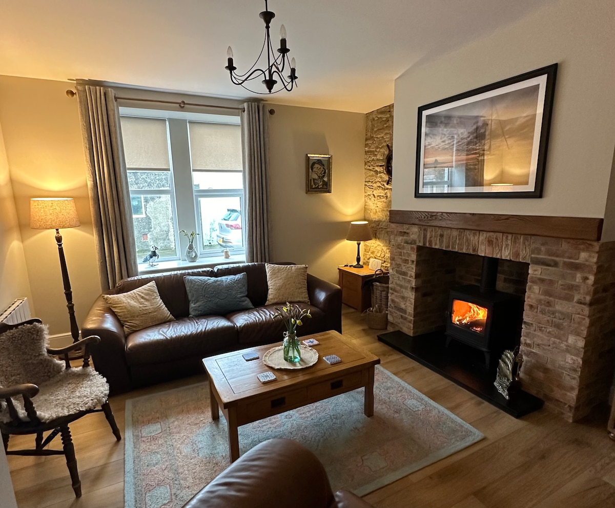 A spacious lounge is presented with a comfortable leather sofa and a wooden coffee table. A log burner is set within a stone fireplace, providing warmth and ambiance. Natural light fills the room through a large window adorned with soft curtains, enhancing the cozy setting.