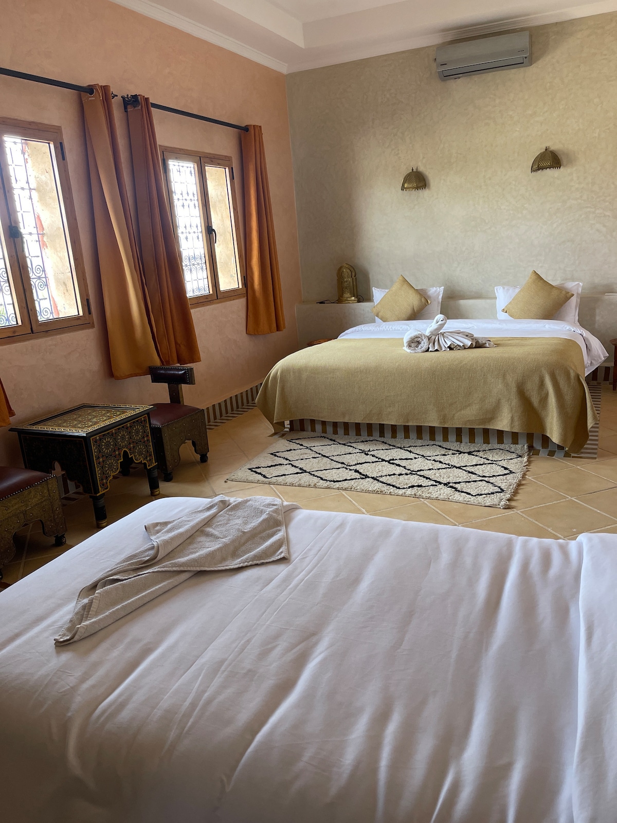 A spacious guest room features two beds adorned with neutral-toned linens, complemented by decorative pillows. Large windows provide natural light, framed by warm curtains. A patterned rug enhances the floor, while a decorative side table adds character to the space.