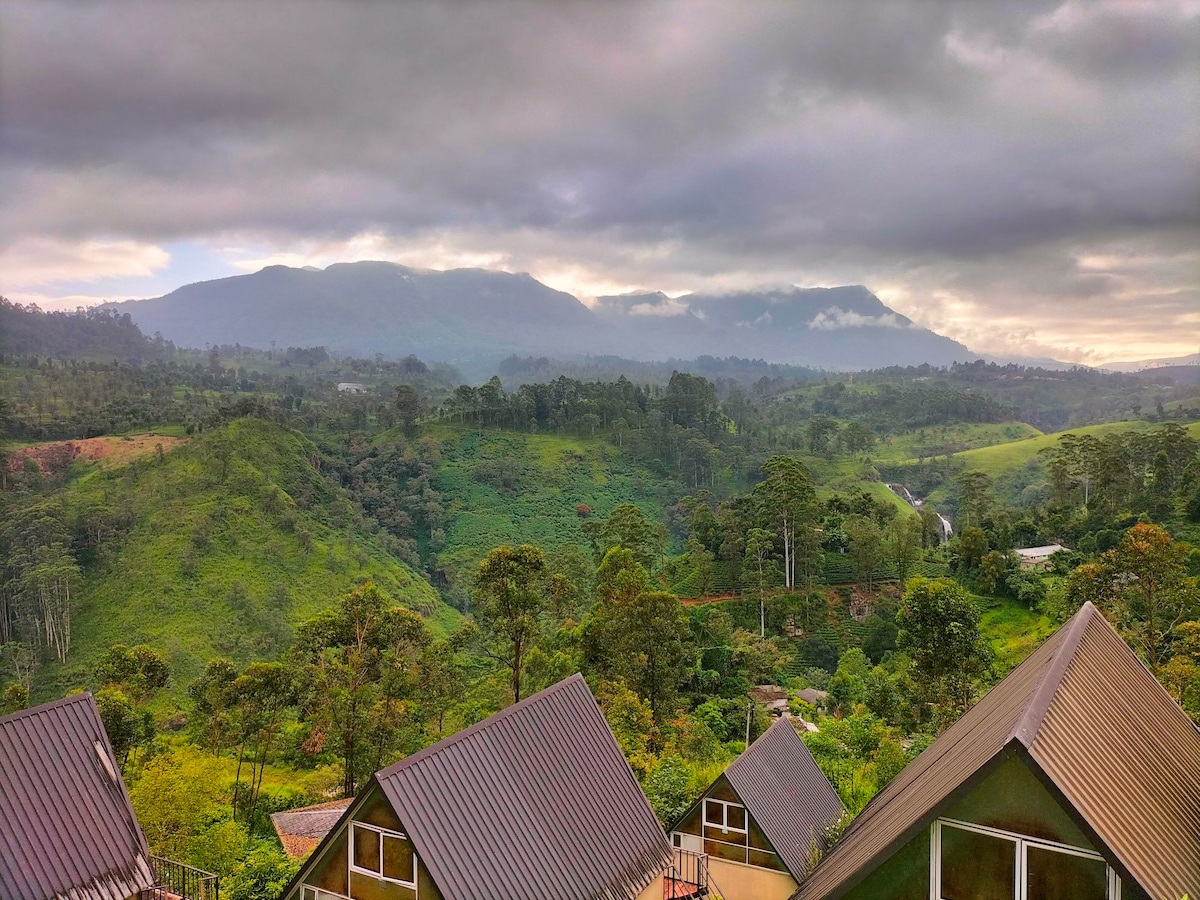A view of rolling green hills and distant mountains is framed by a soft gray sky. Several cabins with sloped roofs are positioned in the foreground, surrounded by lush trees and natural vegetation.