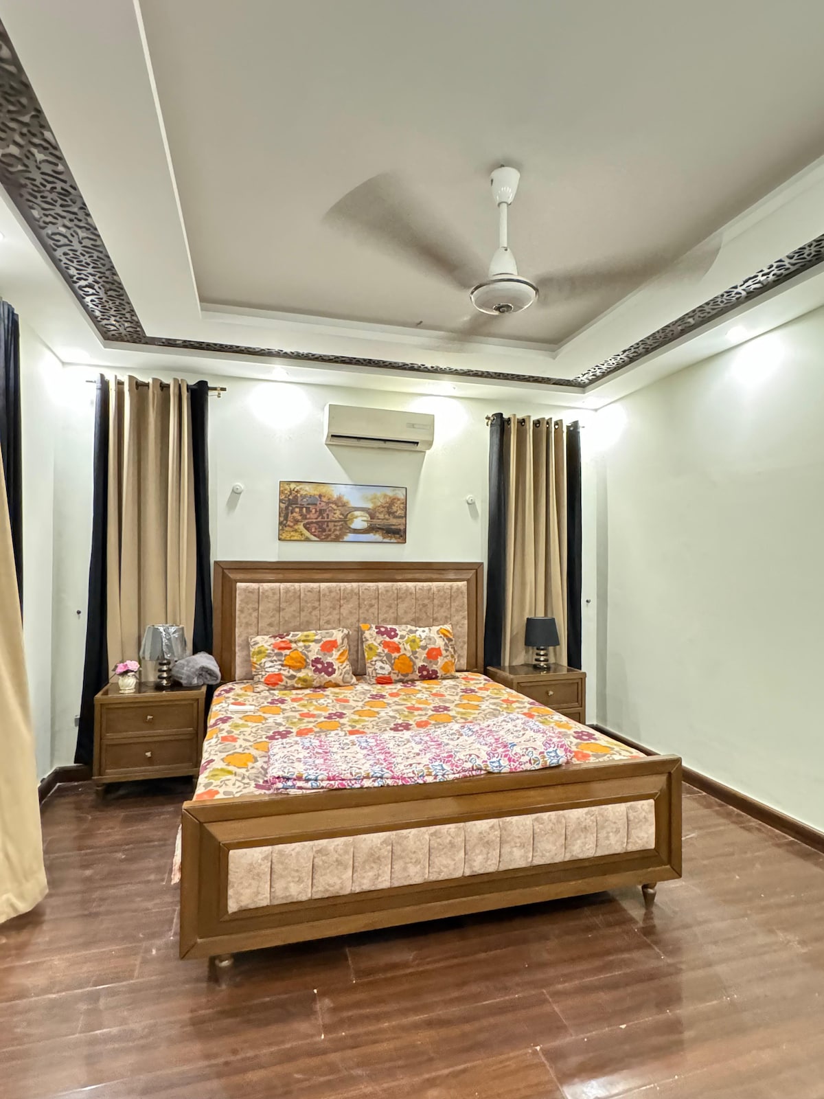 A well-appointed bedroom features a spacious bed adorned with a colorful quilt. Light hardwood flooring is complemented by light-colored walls and decorative ceiling moldings. Black curtains frame a window, while a small bedside table holds a lamp, contributing to a serene atmosphere. An air conditioning unit is visible above.
